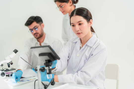 Young Asian woman in lab coat holding a green specimen slide while colleagues work at computers behind, research teamwork in laboratory.