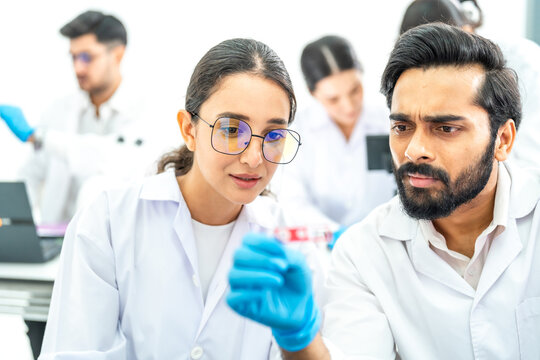 Young Asian woman in lab coat and gloves operating a digital microscope while two colleagues work behind, advanced lab equipment in room.