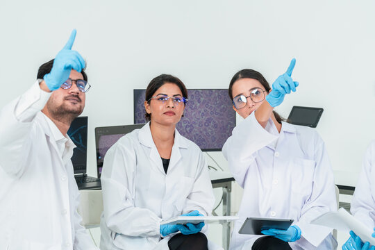 Three researchers in lab coats and gloves pointing and looking up while seated, brainstorming idea in a bright research laboratory with screens.