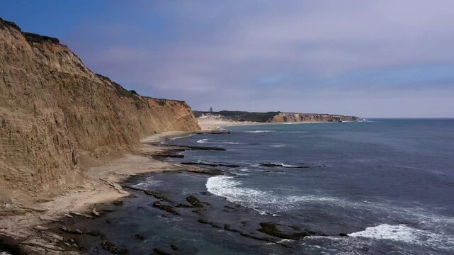 Elevated drone flight tracking south under a bright midday sun, framing rich marine tide pools across the bottom half of the screen below the majestic, jagged and dramatic cliffs.