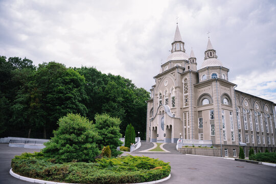 Exterior view of a large, beautiful gray church or cathedral with elegant towers, arched windows, and crosses on the roof, surrounded by green trees, landscaped bushes, 
