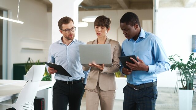 Busy young mixed-race people walking in modern company with gadgets and speaking discussing work project. Group of young business people working in cabinet using laptop and tablet. Corporate concept