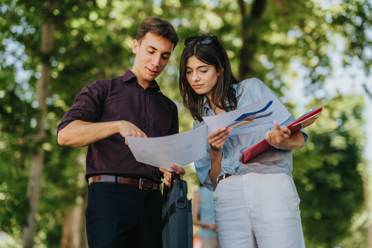 Two young professionals study printed charts and paperwork together while standing outdoors. The colleagues discuss documents and review charts in a relaxed park setting.