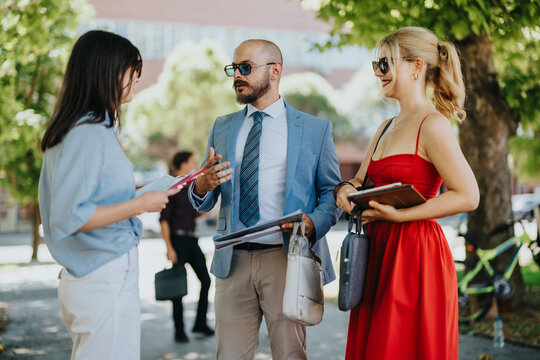 Three colleagues talk and review documents during an outdoor business meeting. A professional discussion takes place between casually dressed coworkers on a sunny campus.