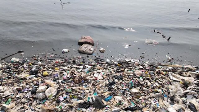 Abandoned cushion and intense plastic waste floating in dark coastal water and muddy shore, demonstrating severe marine littering and ecology problems.