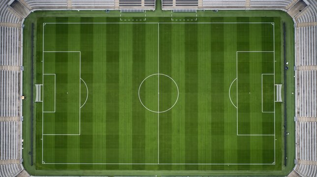 Aerial top-down view of a striped green soccer field inside a stadium, with white lines, center circle, halfway line, penalty areas, goals, and empty stands