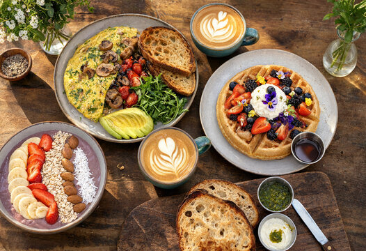 Delicious breakfast spread featuring waffles topped with berries cream avocado toast omelette with mushrooms tomatoes coffee latte art and fresh flowers on rustic wooden table