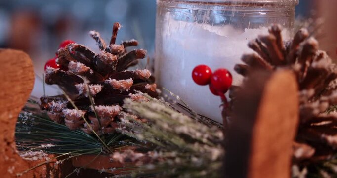 Slow macro movement explores several frosted pinecones bright red berries evergreen branches and a white candle jar while a wooden reindeer shape crosses the soft foreground