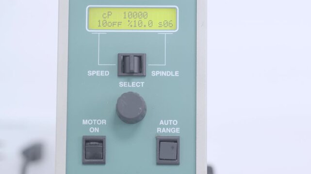 Close-up of a laboratory viscometer: a hand in a blue glove presses the power switch, next to controls for selecting speed, spindle and automatic mode