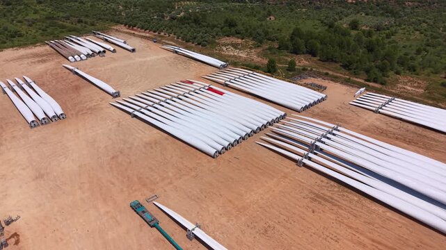 Large wind turbine blades stored in rows on a dry dirt staging area surrounded by sparse vegetation, awaiting installation at a renewable energy construction site