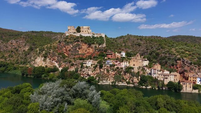 Miravet village and medieval castle perched on rocky cliffs above the Ebro River, Catalonia, Spain, surrounded by green hills and blue sky