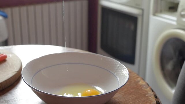 Adult hands cracking chicken eggs into shallow white ceramic bowl with blue rim on worn wooden table, showing fresh yolks, shells and egg white in warm home kitchen preparation, with egg, yolk, bowl