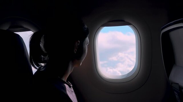Backlit traveler looking out aircraft window at fluffy clouds and blue sky. Passenger sitting by porthole enjoying aerial landscape view during flight. Aviation journey inspiration freedom travel