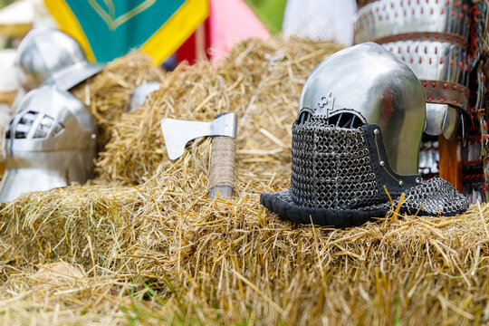 A still life composition featuring historical combat equipment including helmets and an axe, set against a backdrop of hay bales and banners