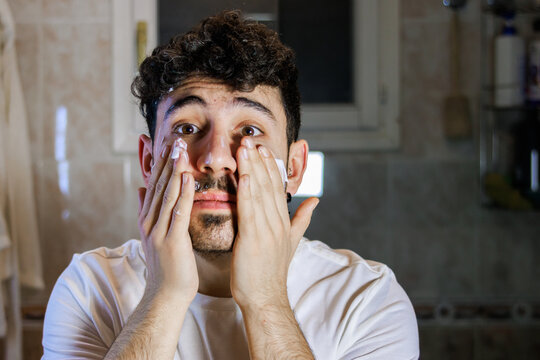 Young man applying facial cream in bathroom for daily skincare routine and grooming