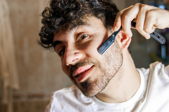 Young man enjoying self-care, shaving beard with traditional straight razor