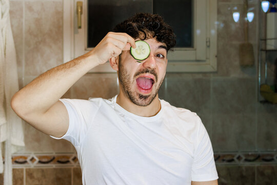 Young man having fun with cucumber slice on eye enjoying self-care routine