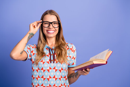 Young woman with glasses smiling holds open book against blue background in a stylish casual student lifestyle portrait