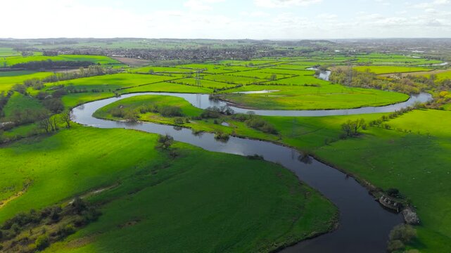Large winding river crossing expansive agricultural land with layered green tones and hedgerow divisions. High altitude aerial view rural fields scattered trees and distant settlements creating depth