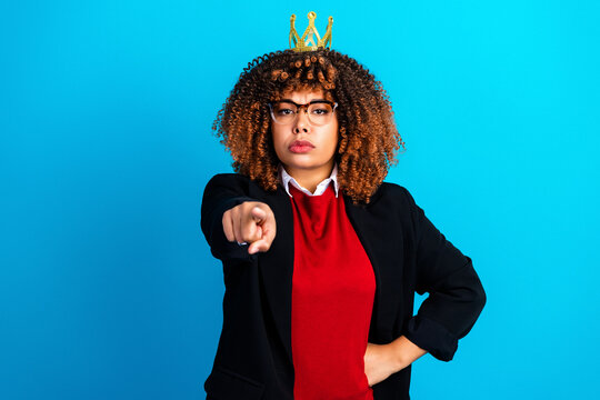 Confident businesswoman wearing a crown points directly at camera, asserting leadership and authority with a serious expression