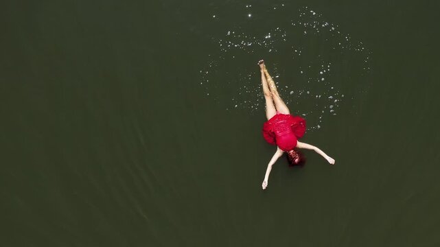  Aerial view of a woman in a red dress swimming in a calm, dark body of water. The sunlight is reflecting on the water