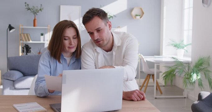 Couple using laptop at home looking through financial documents. At a desk in a bright room, they review bills, compare items, and complete payment with a secure digital app. Online shopping.