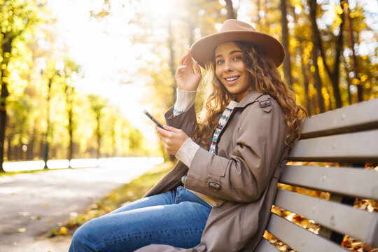 Woman sits on wooden bench, holding phone, surrounded by trees with yellow foliage. Woman in hat and with smartphone enjoys weekend in autumn park at sunset. Concept of seasonality, weekends. 