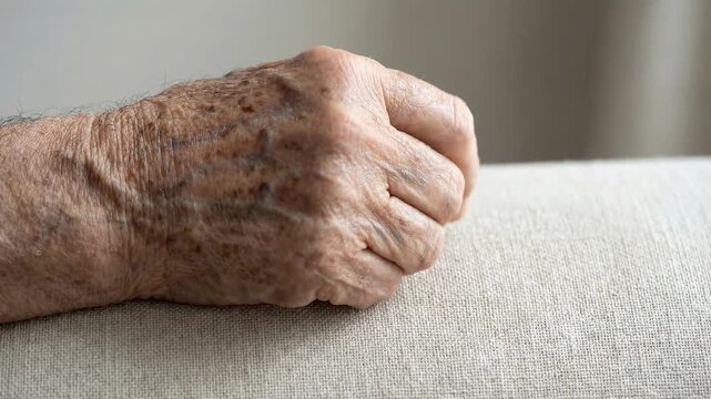 Close-up shows elderly hand palm-side down resting on woven light fabric, fingers slightly curled, knuckles gently bent, visible veins and age spots, with soft diffused light, quiet mood, and