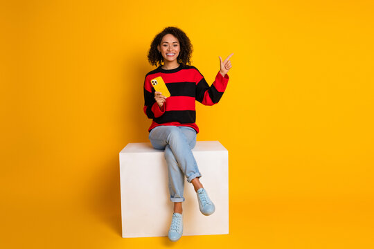 Young woman sits on a white cube smiling in a striped red and black sweater against a yellow background