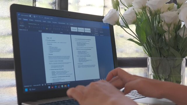 Horizontal video of female hands typing on laptop keyboard with text document on screen beside vase of white flowers