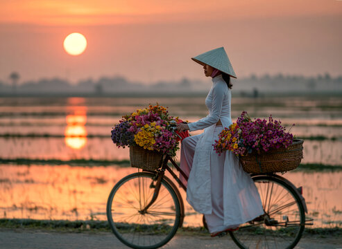 Vietnamese woman wearing a traditional white ao dai and conical hat riding a bicycle full of flowers past a flooded rice paddy at sunset.