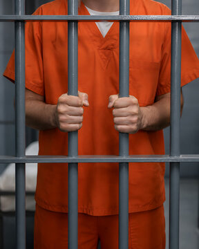 Close up of prison inmate wearing an orange uniform gripping jail cell bars representing incarceration, imprisonment, justice, criminal justice system, and correctional facility