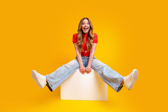 Happy young woman sitting on a white block against a bright yellow background, smiling in a red top, wide jeans and white sneakers