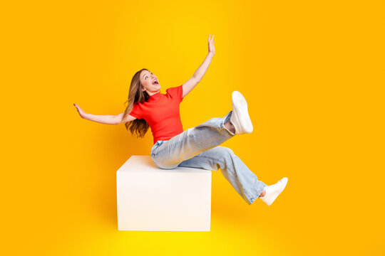Happy young woman in red top and jeans jumping on white cube against bright yellow background