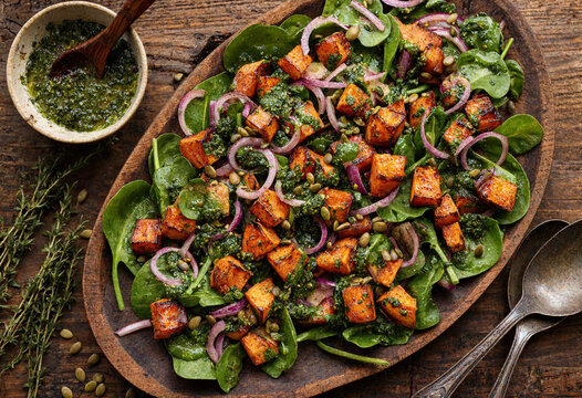 Fresh vibrant spinach salad topped with roasted sweet potato cubes red onion slices pumpkin seeds and herb dressing served on wooden tray