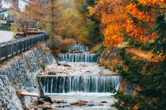 Cascading Waterfall Flow Through Autumn Forest with Vibrant Orange and Golden Foliage Trees in Alpine Valley