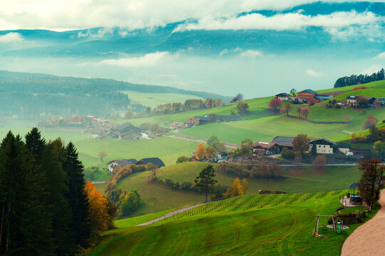 Scenic Alpine Village on Rolling Green Hills with Misty Mountains in Autumn Dolomites Italy