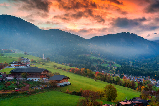 Scenic Alpine Village on Rolling Green Hills with Misty Mountains and Colorful Sky in Autumn Dolomites Italy