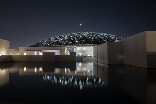 ABU DHABI, UNITED ARAB EMIRATES &ndash; December 01, 2019: Louvre Abu Dhabi Museum illuminated at night, with its iconic dome and architecture reflected in the surrounding water.
