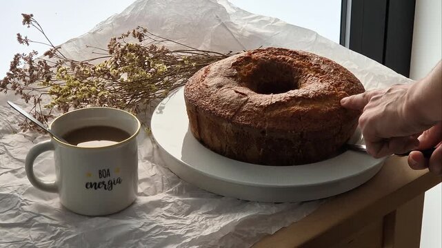 Hand slicing a fresh homemade cake served with coffee. Cozy breakfast or afternoon snack with comforting homemade food in a warm and inviting setting.