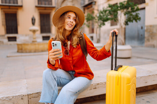 Tourist wearing hat and carrying a brightly colored suitcase sits in town square, holding phone. A young woman with a smartphone is relaxing outdoors. Concept for tourism, weekends, or vacations.