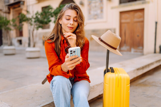 Tourist wearing hat and carrying a brightly colored suitcase sits in town square, holding phone. A young woman with a smartphone is relaxing outdoors. Concept for tourism, weekends, or vacations.