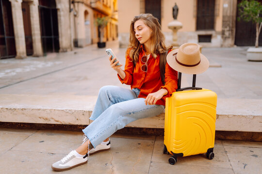 Tourist wearing hat and carrying a brightly colored suitcase sits in town square, holding phone. A young woman with a smartphone is relaxing outdoors. Concept for tourism, weekends, or vacations.