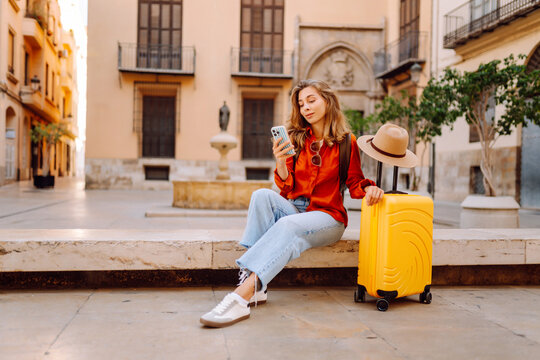Tourist wearing hat and carrying a brightly colored suitcase sits in town square, holding phone. A young woman with a smartphone is relaxing outdoors. Concept for tourism, weekends, or vacations.