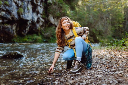 Woman hiker with yellow hiking backpack near mountain river at sunset. Woman enjoys mountain hike and nature in sun's rays. Concept: relaxation, adventure, active lifestyle.