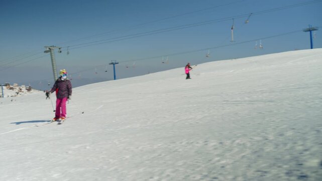 A snowboarder falls on a snowy slope, while a young skier gracefully maneuvers down the mountain. This scene captures the dynamic energy of winter sports at a vibrant ski resort under a clear sky.