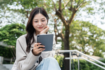 Fototapeta premium Cheerful young Asian businesswoman using digital tablet flexible working life balance in green urban park, modern lifestyle productivity remote work with digital online technology.