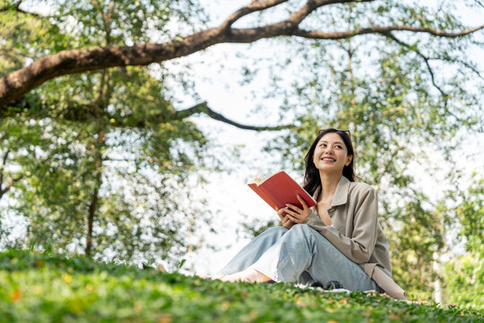 Young asian woman relaxing reading book sitting at public park life style outdoor. Happy female Take a break enjoy time living in the nature. Human and nature together