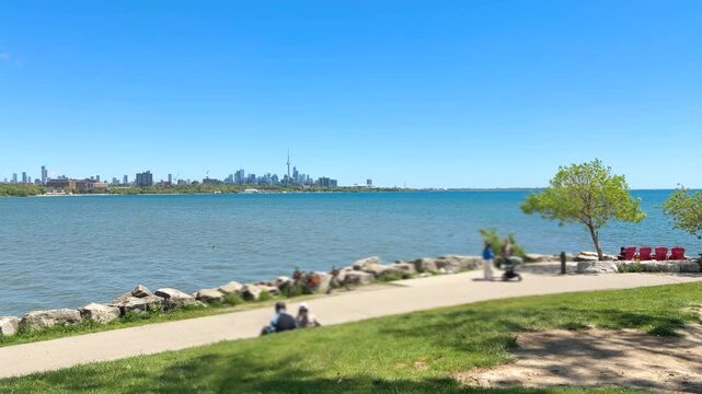 Toronto skyline across Lake Ontario from Sheldon Lookout. People enjoy a sunny summer day relaxing in the green waterfront park. Bicycles rest near the paved promenade under the clear blue sky.