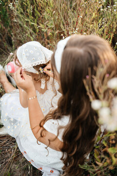 ender moment of a mother caring for her cute little daughter, adjusting her white headscarf and styling small flowers in her hair while sitting together in a wild meadow filled with white flowers. 